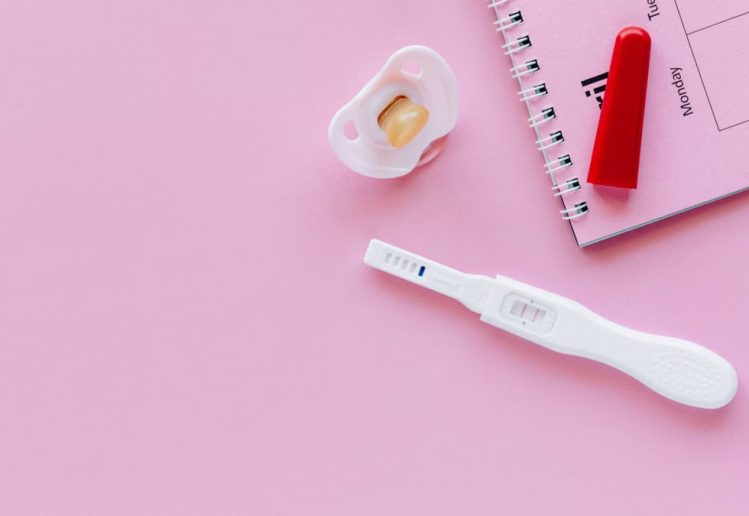 A positive pregnancy test on a pink surface with a pacifier and planner.