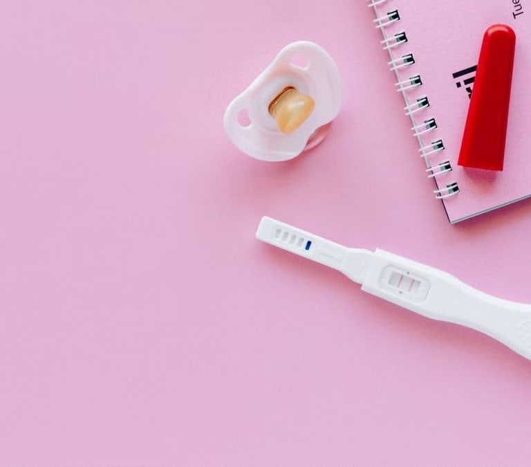 A positive pregnancy test on a pink surface with a pacifier and planner.